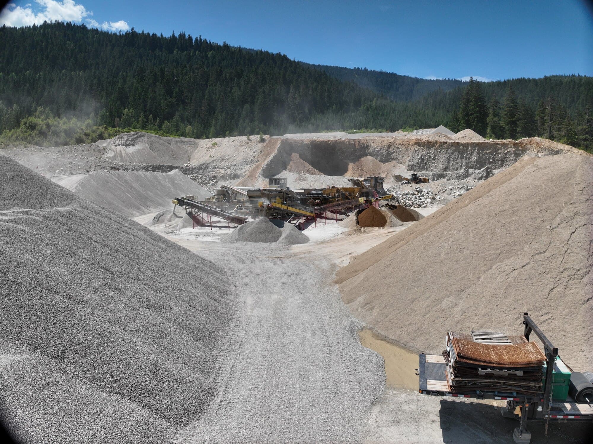 A wide drone view across a working pit, dust drifting off the crushing spread, forested hills on every side.