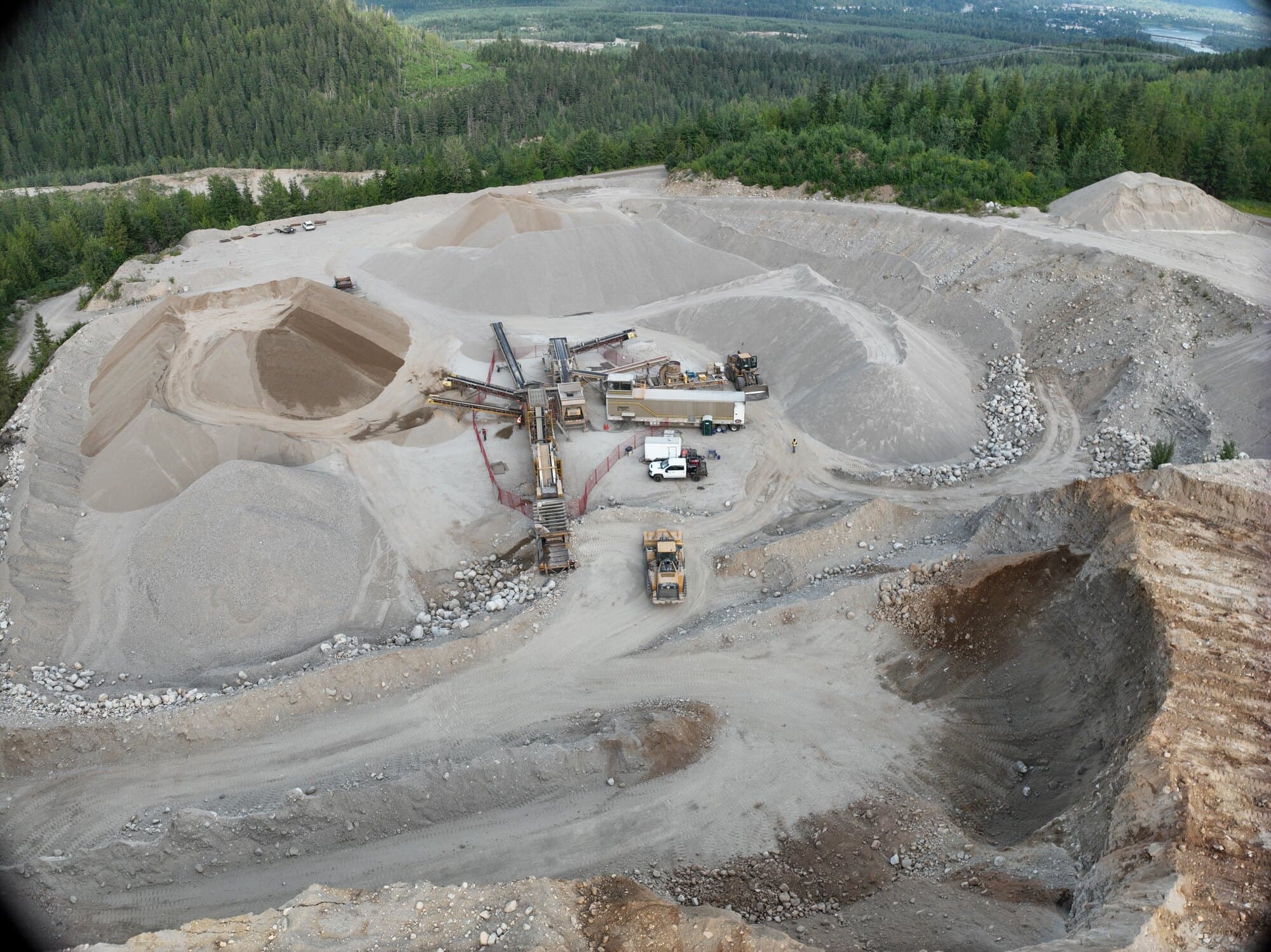A high drone view of a whole working pit: the crushing spread at its centre, stockpiles and benches around it, a river valley beyond.