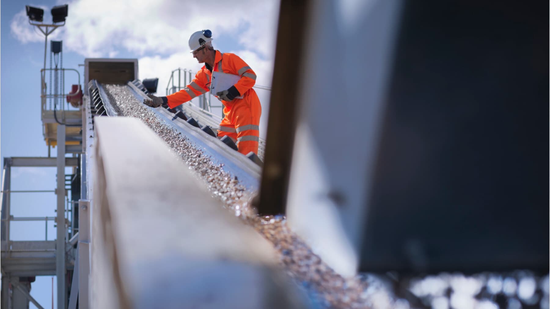 A worker in hi-vis coveralls and a hard hat checking crushed aggregate coming off a conveyor against a clipboard.