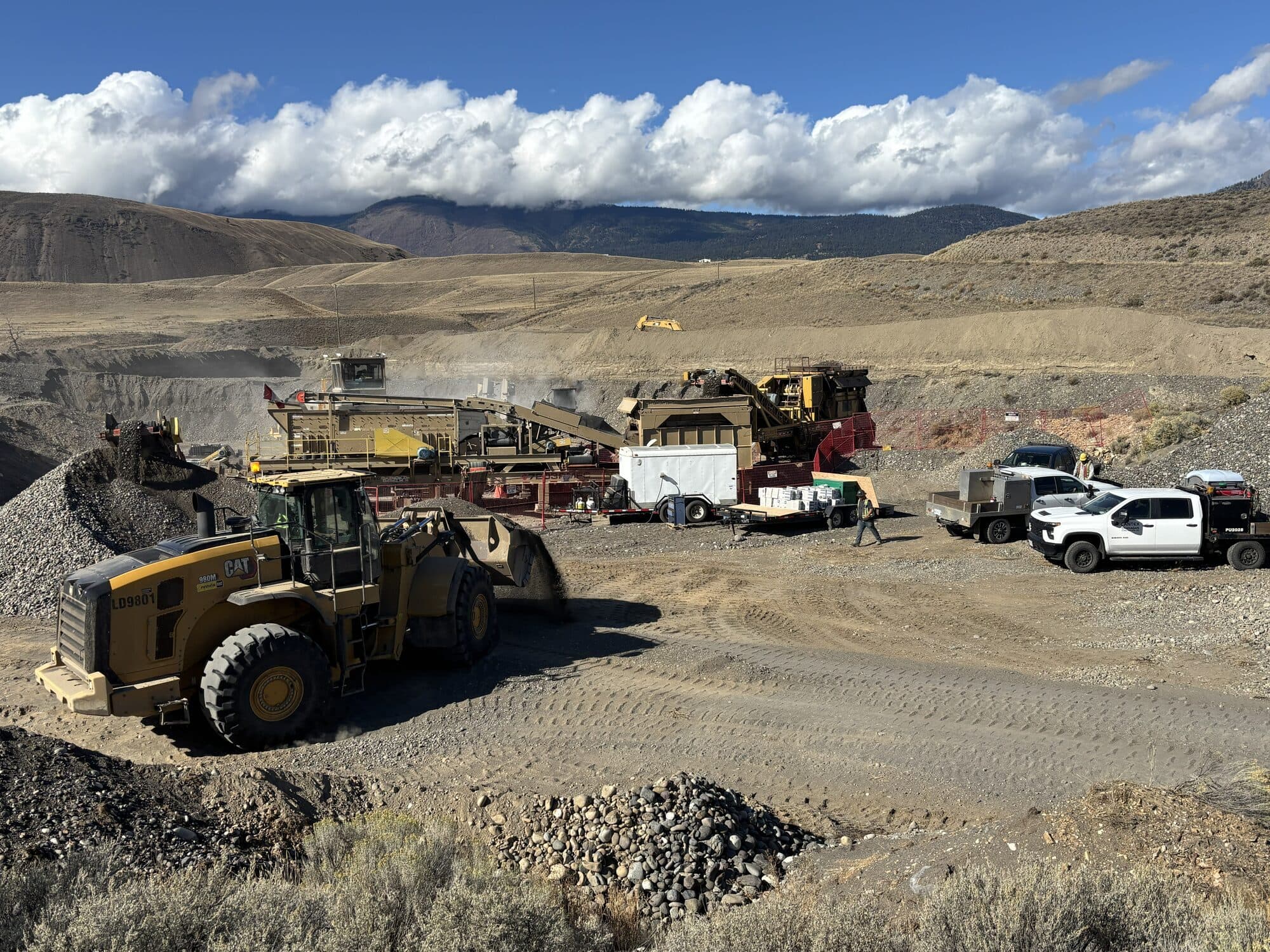 A wide Cantex crushing operation: a wheel loader in the foreground, crew and trucks on site, mountains behind.
