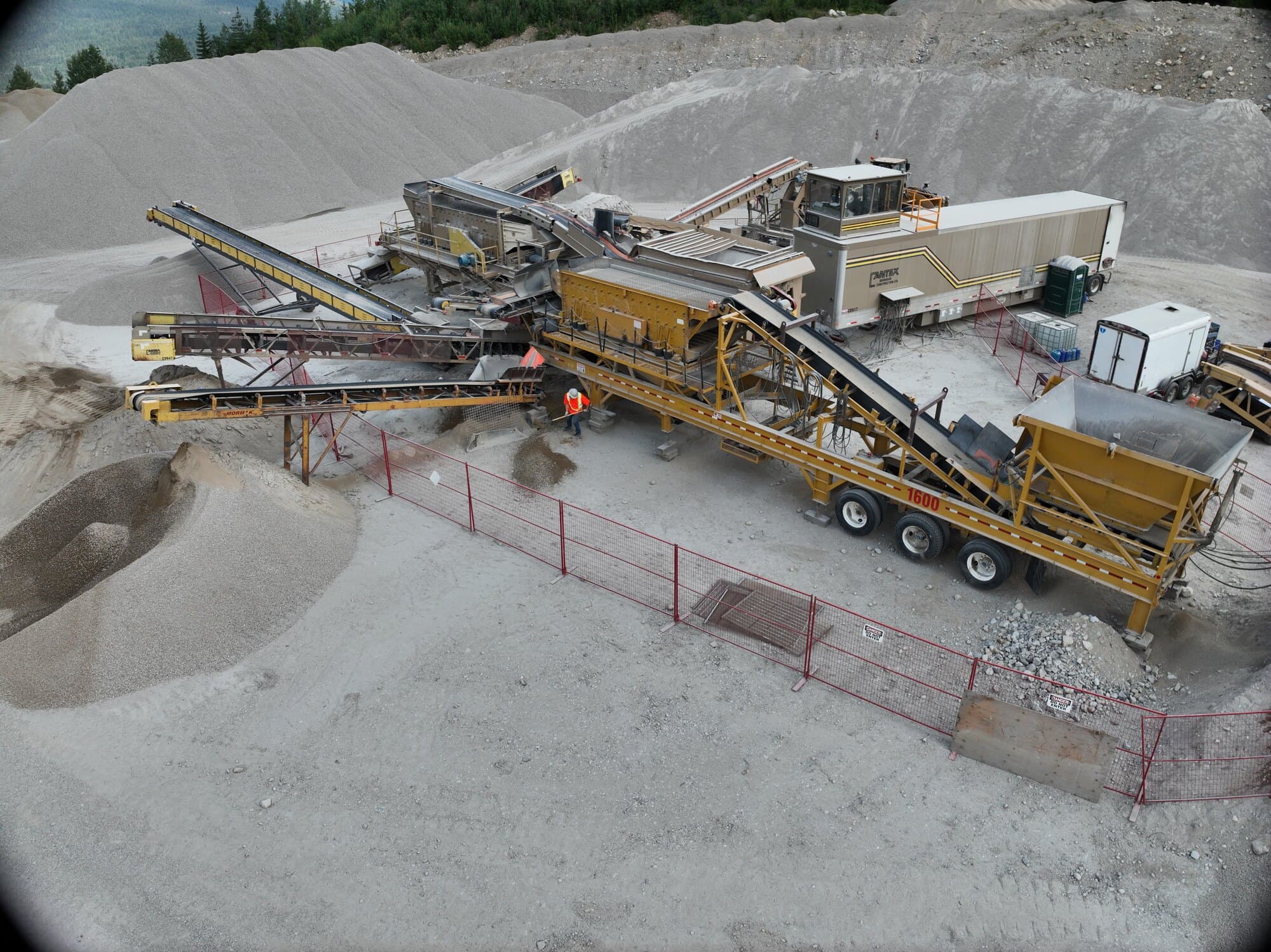 A Cantex operator in hi-vis walking the length of a running crushing spread, stockpiles and forested hills behind.