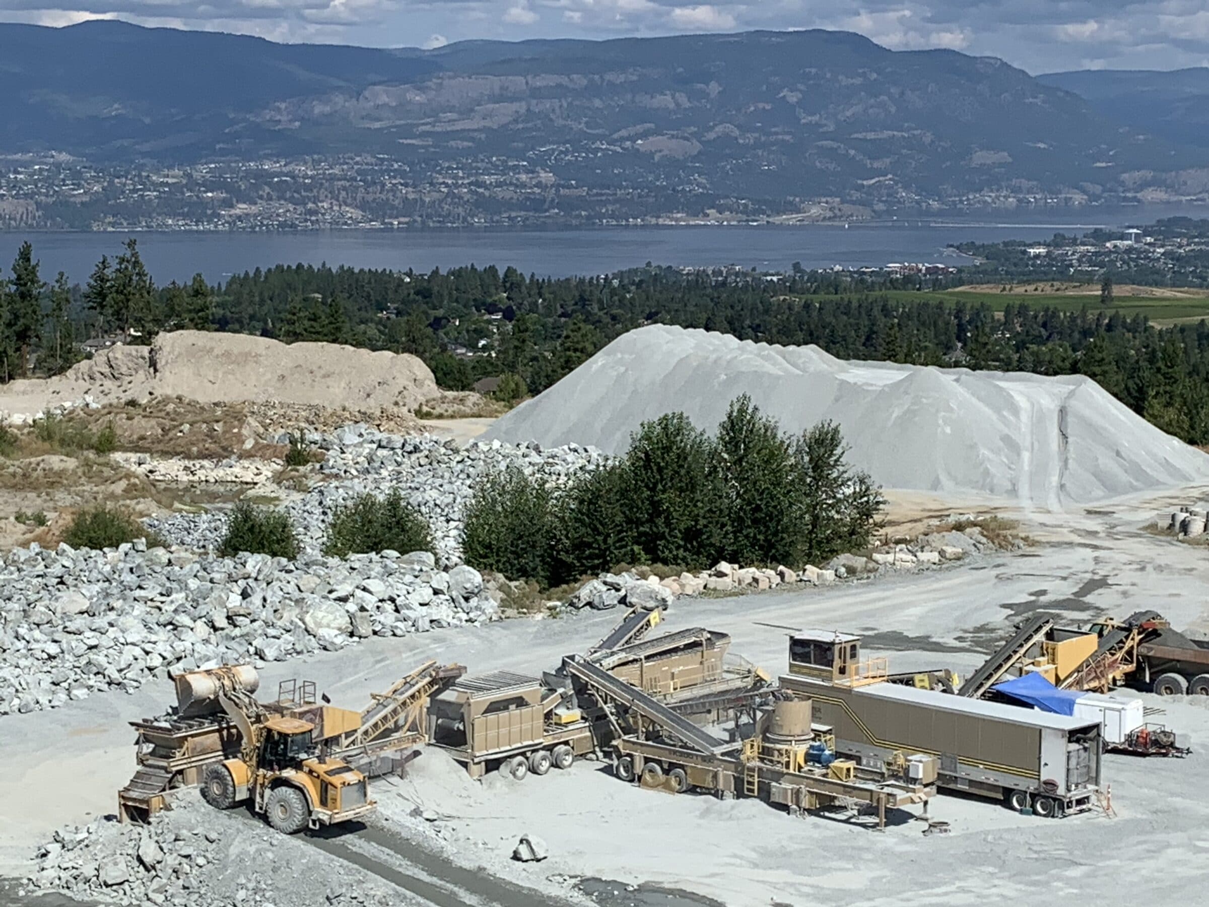 A Cantex crushing spread and wheel loader working a quarry above a lake, with a valley and mountains beyond.