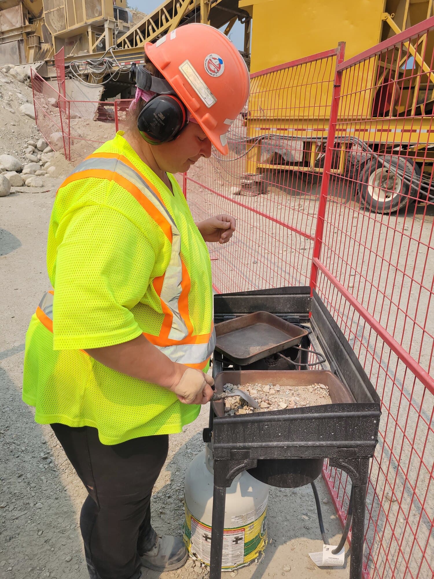 A Cantex worker in hi-vis and hearing protection splitting a crushed-aggregate sample into trays at an on-site sample station, with the plant behind.