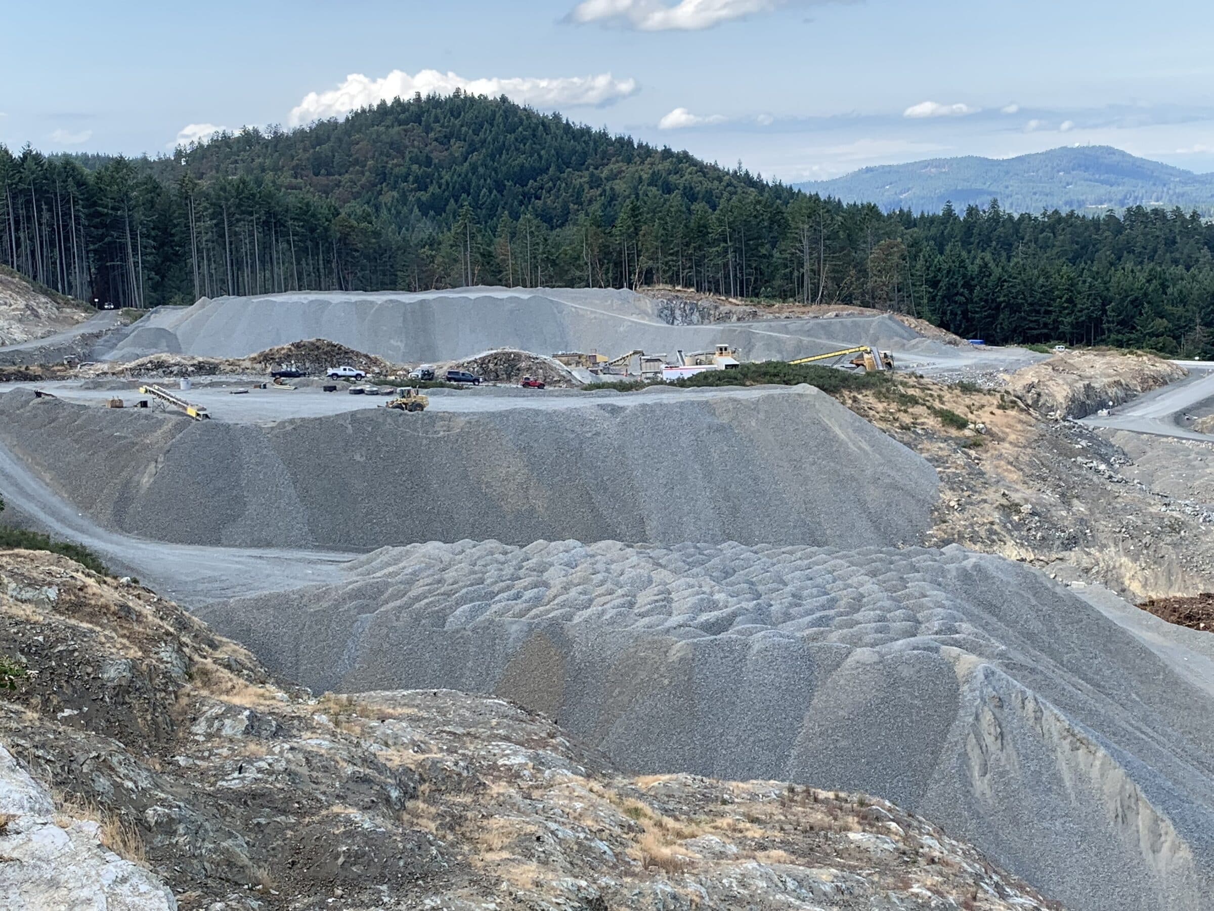 A large quarry face and graded stockpiles with a crushing spread working along the bench, coastal forest behind.